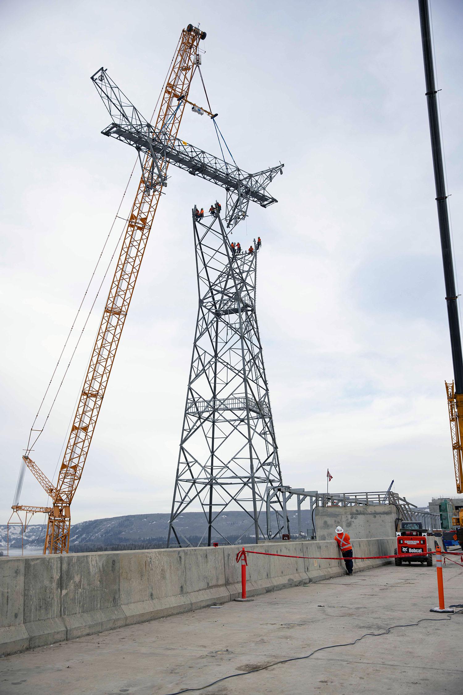 A spreader bar is used to keep the head section of the transmission tower level as it is lowered onto four connection points. Two crew members wait at each point to bolt the sections.