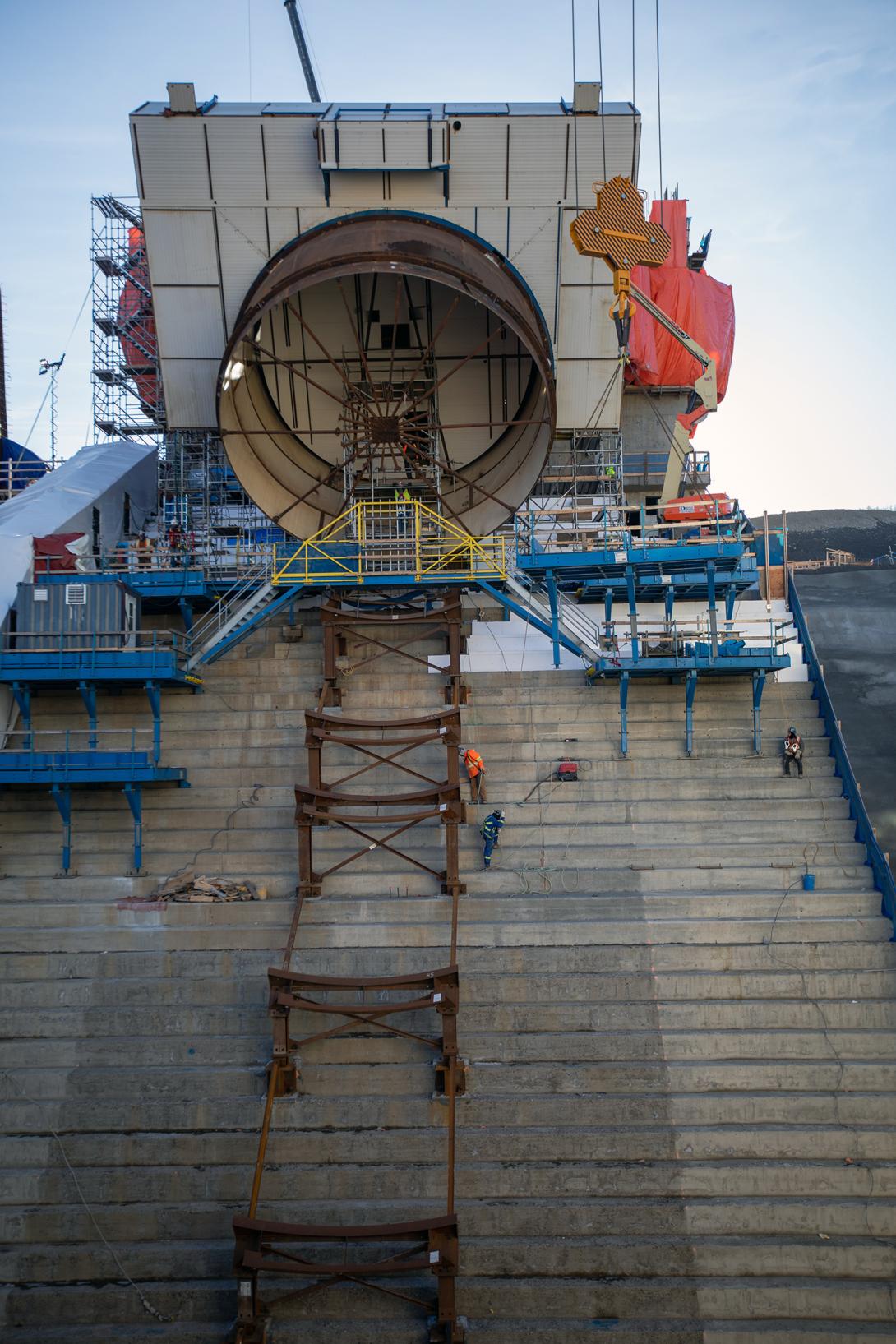 The square, white collar around the penstock is an external scaffolding to protect welders while they join the seams.  More than 40% of the welding is complete and 13 of 18 pieces needed for this penstock are on site and in position. | October 2019