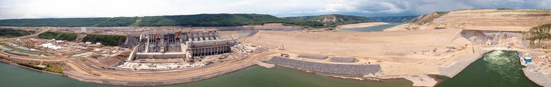 Panorama of spillways, powerhouse and generating station, completed earthfill dam, and diversion tunnel outlet portals. 