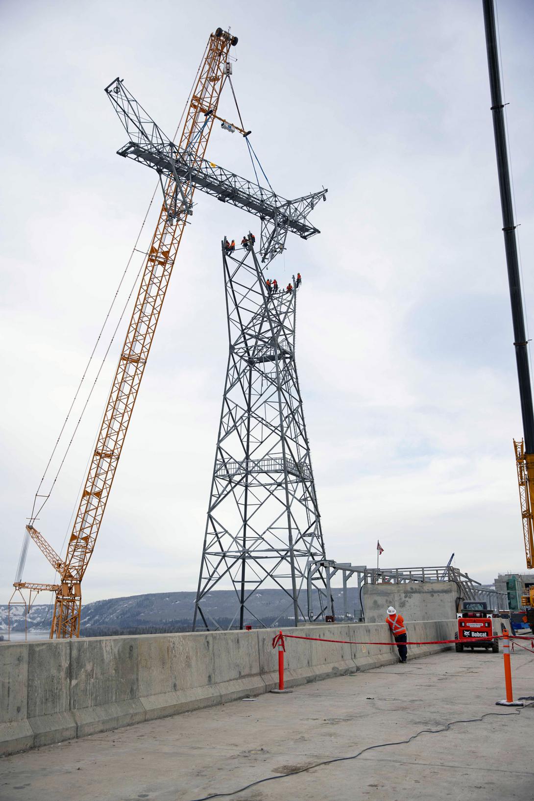 A spreader bar is used to keep the head section of the transmission tower level as it is lowered onto four connection points. Two crew members wait at each point to bolt the sections. 