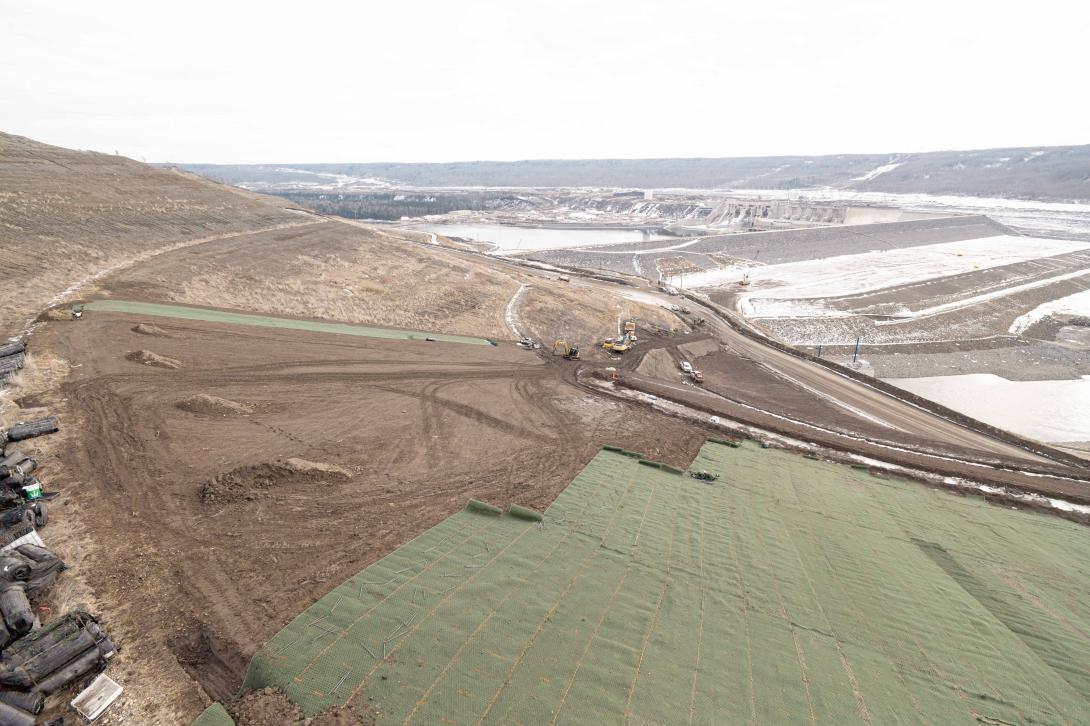The green synthetic matting is rolled over seeded ground and anchored with long pins. The mesh allows real grass to grow through it on the hills above the Site C dam. The synthetic blanket prevents soil from sliding off the slope.