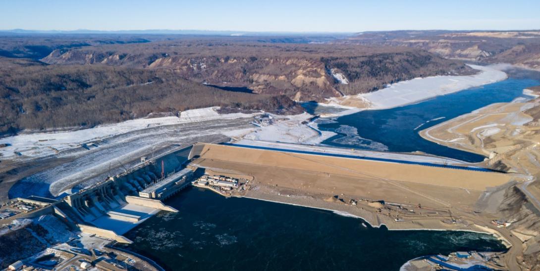 Facing the downstream side of the completed Site C dam, with the approach channel behind the spillways, penstocks, and the powerhouse and operations building at left.