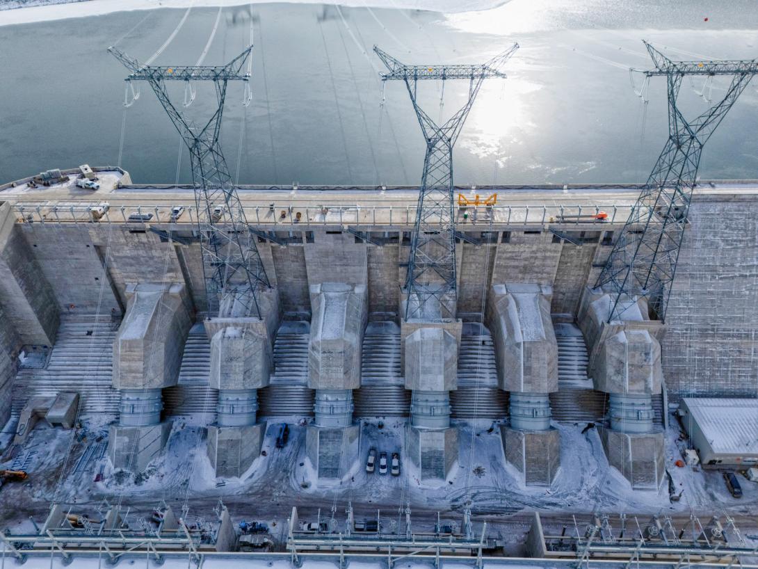 Water flows from the approach channel, via the intakes, through the penstocks and into the powerhouse. From the powerhouse, electricity passes through the sets of transformers (bottom of photo) and is transmitted to the substation by the lines atop the transmission towers. | March 2025