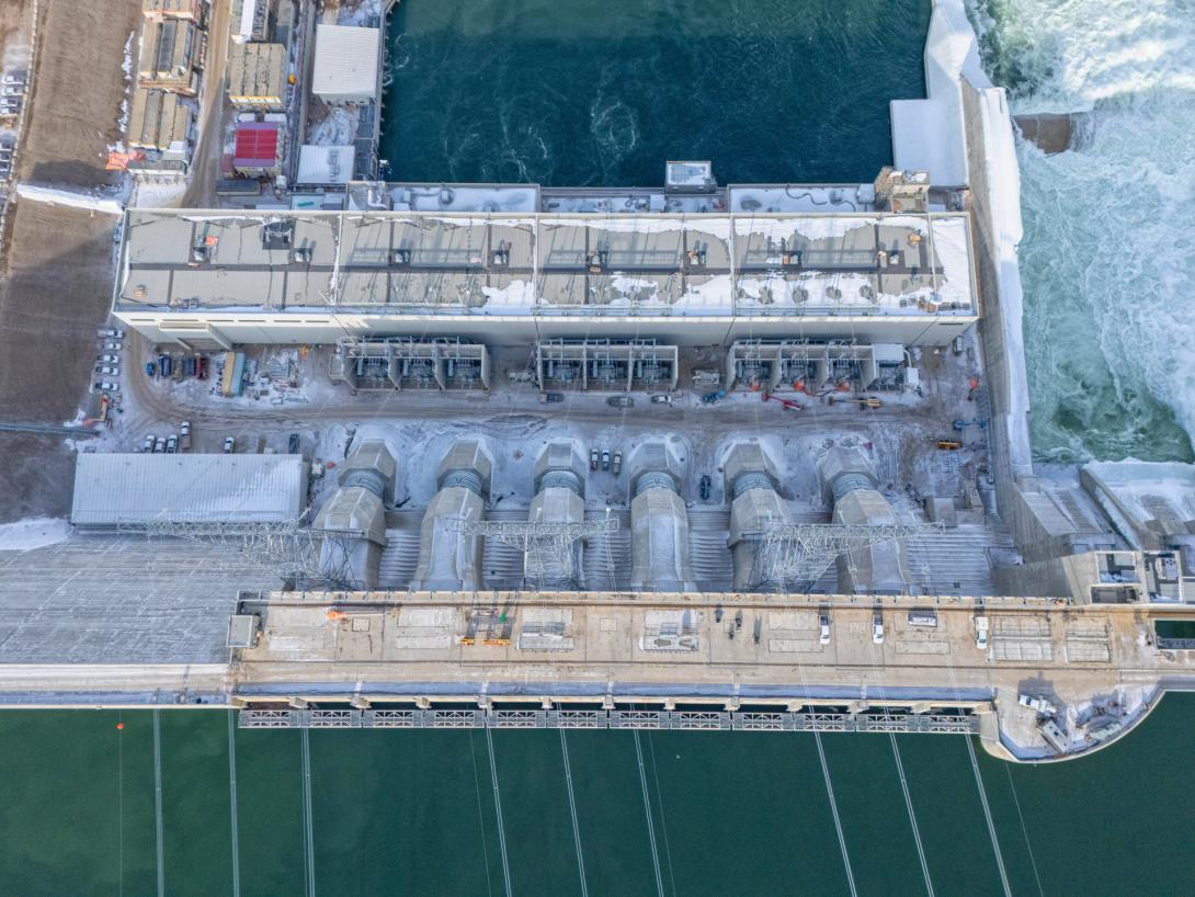Water from the approach channel flows via the intakes into the six penstocks. The penstocks convey water into the powerhouse to drive the turbines and generators. Electricity is generated at 13.8 kilovolts and stepped up to 500 kilovolts before feeding into the transmission lines visible at the bottom of the photo. After passing through the turbines, all the water flows out of the powerhouse into the Peace River. | March 2025