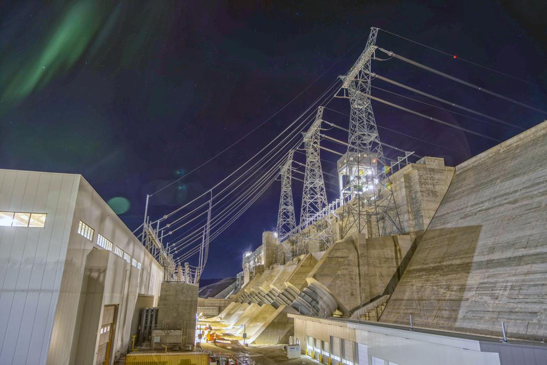 The rear of the powerhouse in the transformer yard, with penstocks, overhead transmission lines and towers. | April 2025
