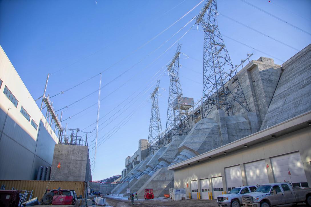 The transformer yard behind the powerhouse. Water travels from the approach channel into the penstocks at right and down 80 metres to the lower levels of the powerhouse. Water spins the turbines to generate electricity. The power is routed out to the transformers at left and then carried up and over the approach channel on lines supported by the transmission towers above. | March 2025